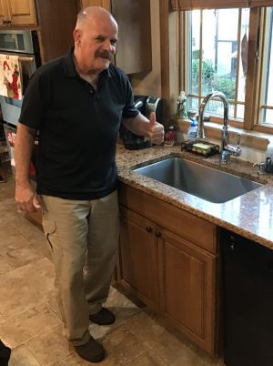 An older man stands in a kitchen next to a sink, giving a thumbs-up gesture.