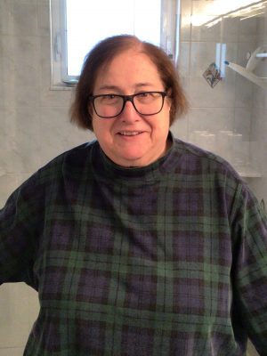 A person with short brown hair and glasses stands indoors, wearing a green and black plaid shirt. White tiled walls and a window are visible in the background.