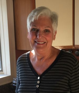 Older woman with short white hair and a striped shirt stands smiling in a kitchen with light from a nearby window illuminating her face.