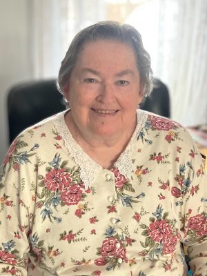 An older woman with short gray hair is sitting and smiling. She is wearing a cream-colored floral top with a white lace collar. The background is softly lit with sheer curtains.