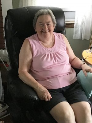 An older woman with short gray hair, wearing a pink sleeveless top and black shorts, sits and smiles on a black recliner chair in a living room.