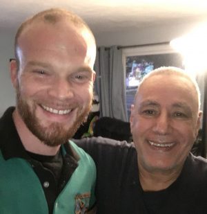 Two men are standing indoors, smiling at the camera. One is wearing a green and black collared shirt, and the other is wearing a dark shirt. A window and curtains are visible in the background.