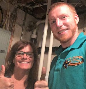 Two people standing indoors give thumbs up and smile at the camera; one wears glasses and the other wears a teal polo shirt with a logo.