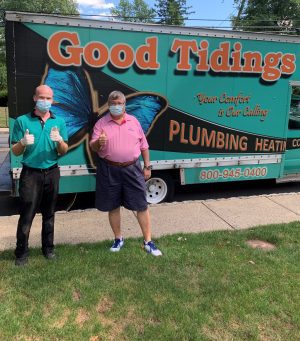 Two men wearing masks stand on grass giving thumbs up in front of a truck with "Good Tidings Plumbing Heating" and a butterfly graphic on the side.