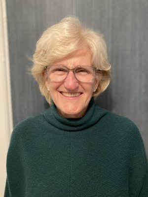 An older woman with short, white hair and glasses smiles at the camera, wearing a dark green turtleneck sweater. The background is plain and gray.