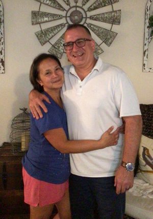 A man and a woman stand indoors, smiling and posing together with their arms around each other. There is wall decor and furniture in the background.