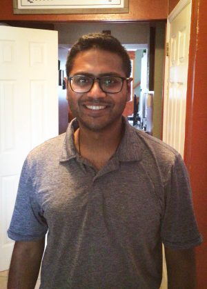A man wearing glasses and a gray polo shirt stands indoors, smiling at the camera. A white door and hallway are visible in the background.