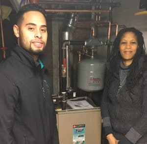 Two people stand next to a residential heating system with visible pipes and a pressure tank in a utility room.