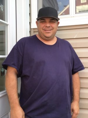 A man wearing a black cap and dark blue t-shirt stands outside near a beige vinyl siding wall and a white door, smiling at the camera.