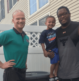 Three people posed outdoors: a man in a green shirt, a man in a black shirt holding a smiling young boy, with a building and white fence in the background.