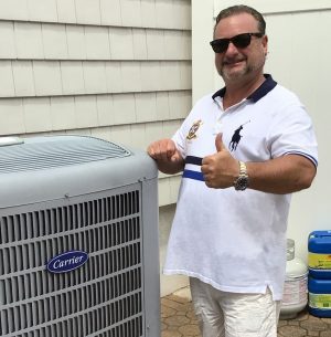 A man wearing sunglasses and a white polo shirt gives a thumbs up next to a Carrier air conditioning unit outdoors.