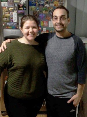 Two people stand side by side indoors, smiling at the camera. The woman wears a green sweater, and the man wears a gray shirt. A fridge with magnets is visible in the background.