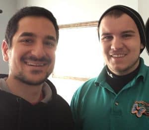 Two men are standing indoors, smiling at the camera; one wears a black hoodie, the other a green polo shirt and a beanie.