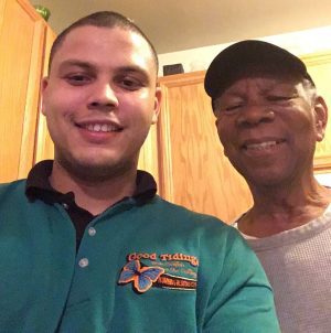 Two men standing side by side, smiling at the camera in a kitchen. One wears a green collared shirt and the other wears a light sweater and a black cap.