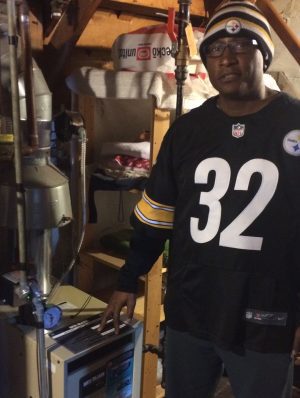 A man wearing a Steelers jersey and hat stands next to a furnace in a basement, with his hand resting on the unit.
