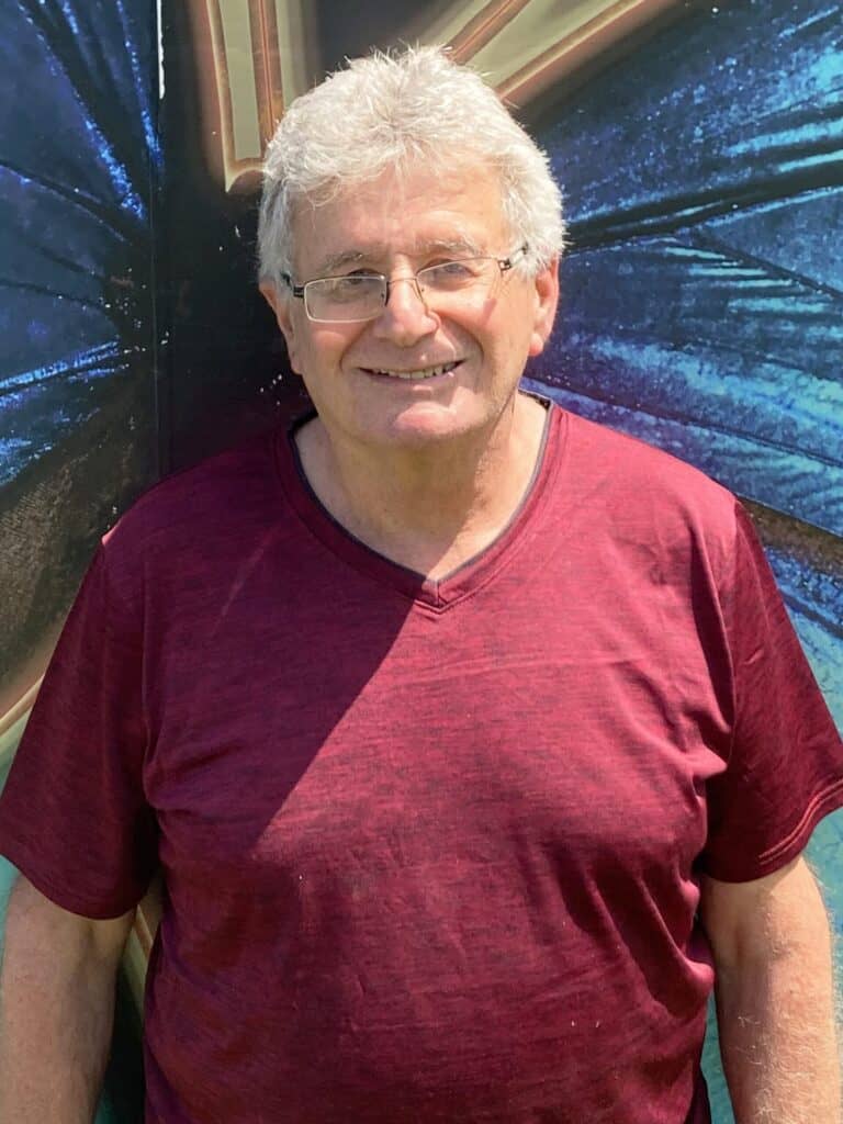An older man with gray hair and glasses, wearing a maroon shirt, stands and smiles in front of a colorful, abstract background.