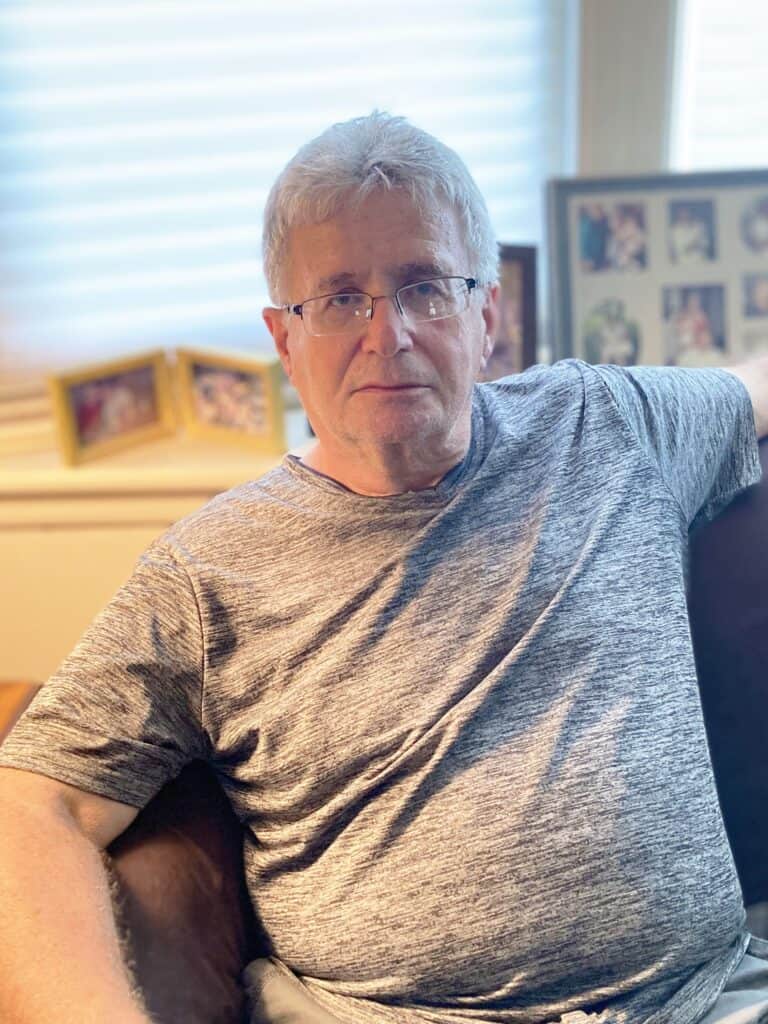 An older man with gray hair and glasses sits on a couch in a well-lit room, with family photos displayed on the windowsill behind him.