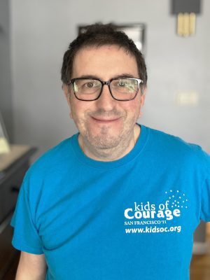 A man with short dark hair and glasses smiles at the camera, wearing a blue "Kids of Courage" San Francisco t-shirt.