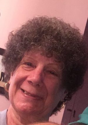 An older woman with short, curly gray hair and gold hoop earrings smiling indoors with a pink wall in the background.