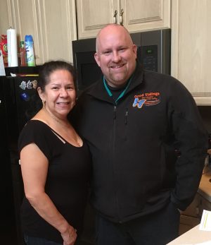A woman and a man stand side by side in a kitchen, both smiling at the camera. The man is wearing a black jacket with an embroidered logo.