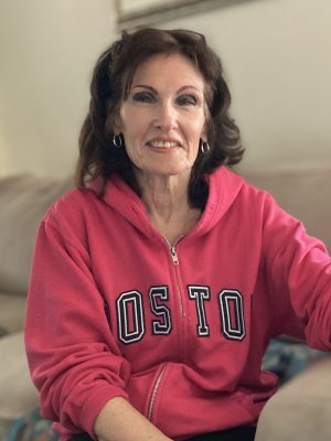 A woman with brown hair sits on a couch, wearing a pink zip-up hoodie with "BOSTON" written on the front.