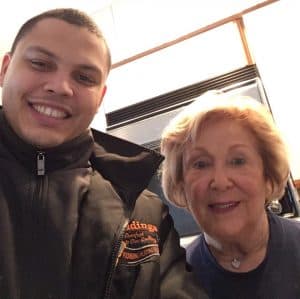 A young man and an older woman smile for a selfie indoors, standing close together in front of a kitchen oven.