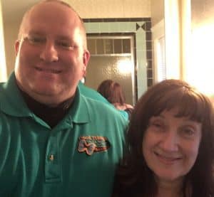A man in a teal collared shirt stands next to a woman with dark hair, both smiling in what appears to be a bathroom with a tiled shower in the background.