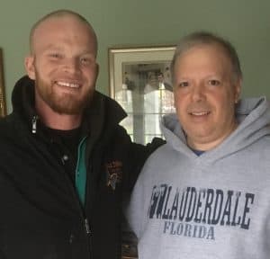 Two men stand indoors smiling at the camera; one wears a black jacket, and the other wears a gray "Ft. Lauderdale Florida" sweatshirt.