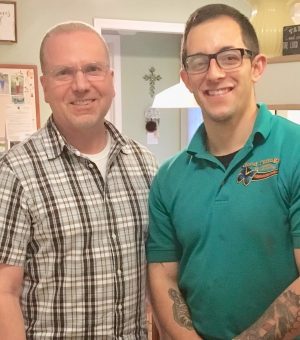 Two men stand indoors, smiling at the camera. One wears a plaid shirt and glasses; the other wears a teal polo shirt and has visible tattoos on his forearms.