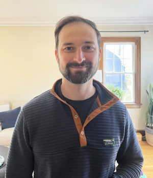 A man with short brown hair and a beard stands indoors, wearing a navy pullover with a brown-trimmed collar. There is a window and houseplant in the background.