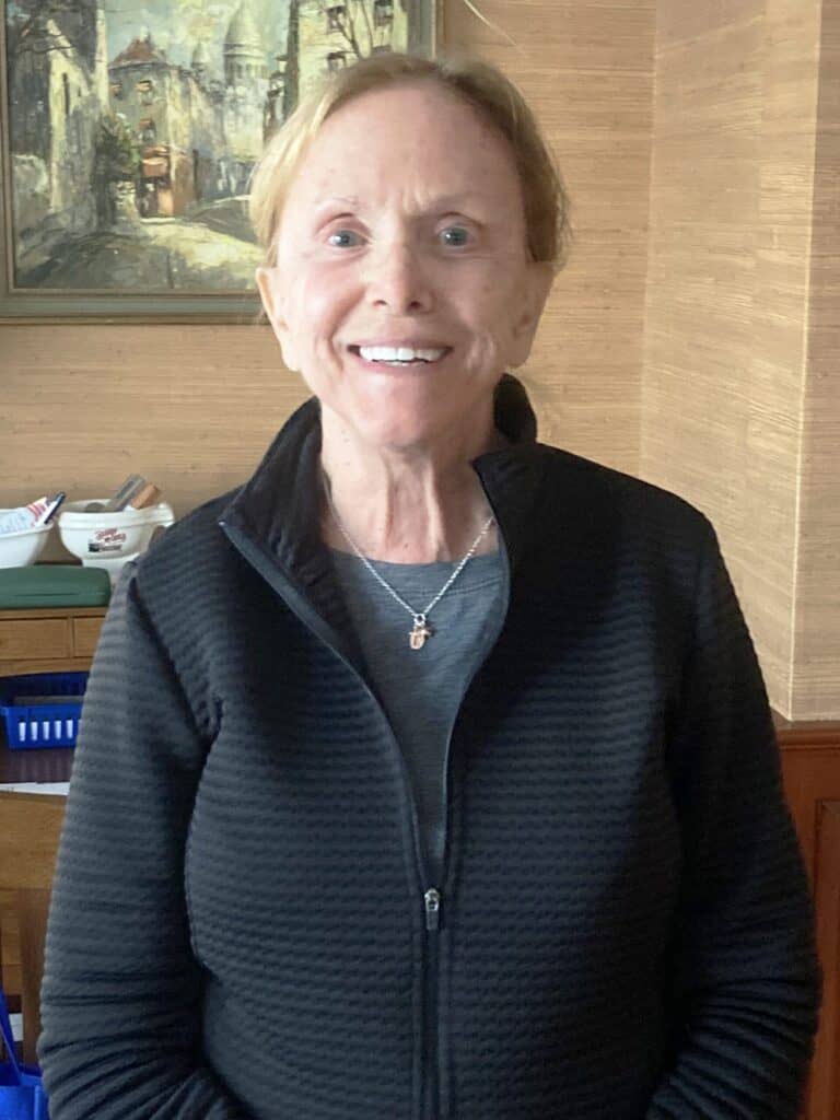 A woman with short light hair, wearing a black zip-up jacket and a necklace, stands indoors and smiles at the camera. A painting and household items are visible in the background.