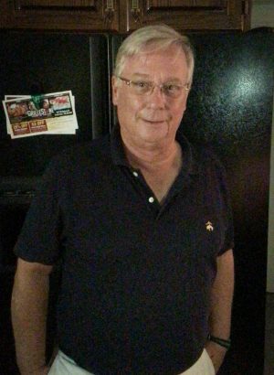 An older man with short gray hair and glasses stands in front of a black refrigerator, wearing a dark polo shirt.