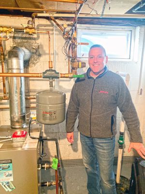 A man stands beside a home boiler system with exposed pipes in a basement, next to a small window.