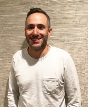 A man with short hair and a beard is standing in front of a textured beige wall, wearing a white long-sleeved shirt with a chest pocket.