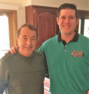 Two men stand indoors, smiling at the camera; one wears a green sweater and the other a teal polo shirt with a logo.