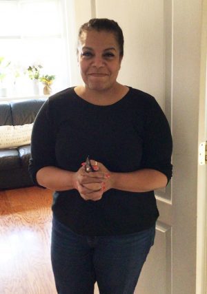 A woman in a black shirt and jeans stands indoors, smiling, with her hands clasped in front of her. A couch and window are visible in the background.