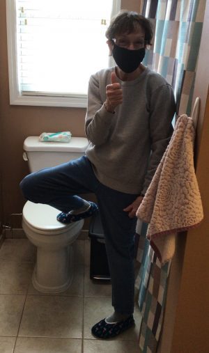Person wearing a mask and casual clothes stands in a bathroom, giving a thumbs-up with one foot resting on a closed toilet lid.