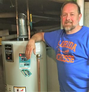 A man in a blue "Florida Lacrosse" T-shirt stands next to a water heater in a basement, with his arm resting on the heater.