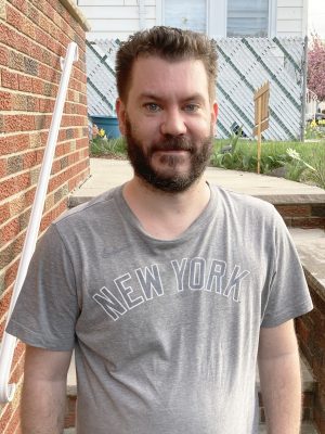 A man with short brown hair and a beard wearing a gray "New York" t-shirt stands outside near brick steps and a handrail.