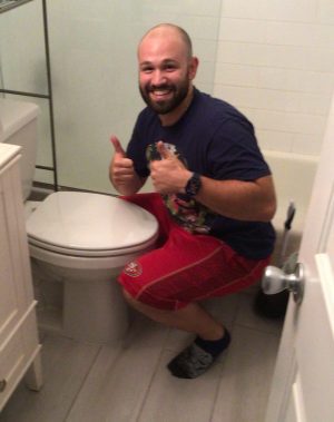 A man in a navy shirt and red shorts crouches next to a closed toilet, smiling and giving two thumbs up in a bathroom.