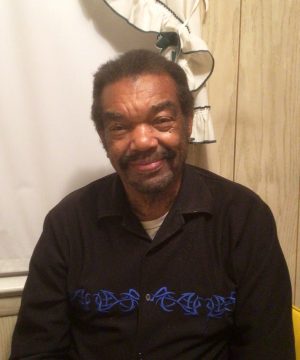A man with short dark hair and a beard sits indoors, wearing a black shirt with blue embroidery, in front of a curtain and wood-paneled wall.