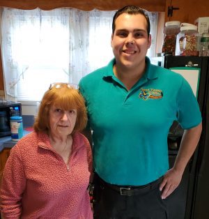 An older woman in a pink pullover stands next to a younger man in a teal polo shirt inside a kitchen with light coming through a window behind them.