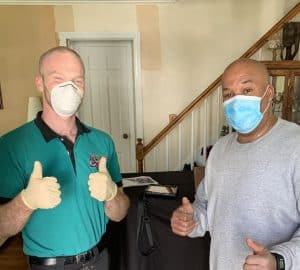 Two men indoors wearing face masks; one in a green shirt and gloves gives two thumbs up, the other in a gray shirt stands beside him near a staircase.