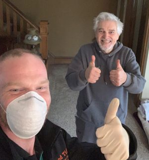 Two men indoors: one wearing a face mask and glove giving a thumbs up, the other smiling and giving two thumbs up.