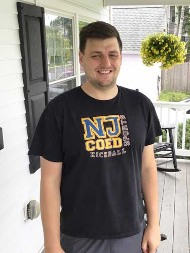 Man standing on a porch wearing a black "NJ Coed Kickball Sports" t-shirt, with a hanging basket of yellow flowers in the background.