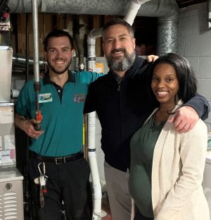 Three adults stand together in a basement next to HVAC equipment; one is wearing a work uniform and holding tools, while the other two smile at the camera.