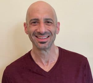 A bald man with a trimmed goatee smiles at the camera, wearing a maroon V-neck shirt against a plain light-colored background.