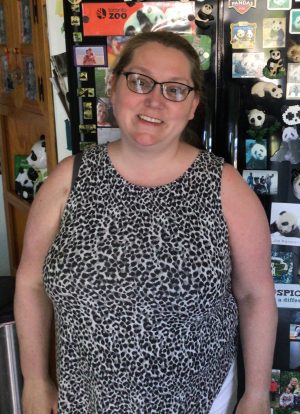 A woman wearing glasses and a sleeveless leopard print top stands indoors in front of a refrigerator decorated with panda magnets and photos.