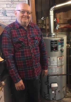 An older man wearing glasses and a plaid shirt stands next to a water heater in a basement or utility room.