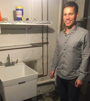 A man stands next to a utility sink in a basement with exposed pipes and a shelf holding cleaning supplies.